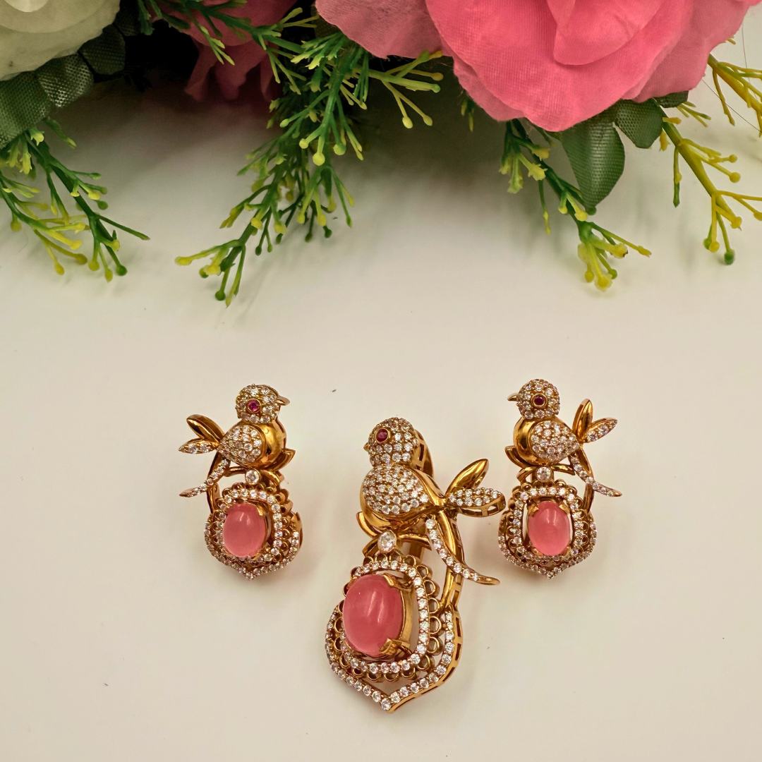 Gold bird-shaped earrings with pink stones on a white background with greenery.